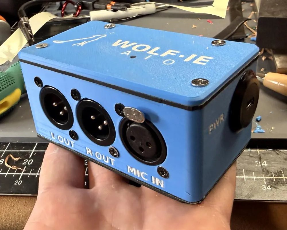 The Wolf-ie AIO box held in one hand, front face showing L OUT, R OUT and MIC IN XLR connectors
